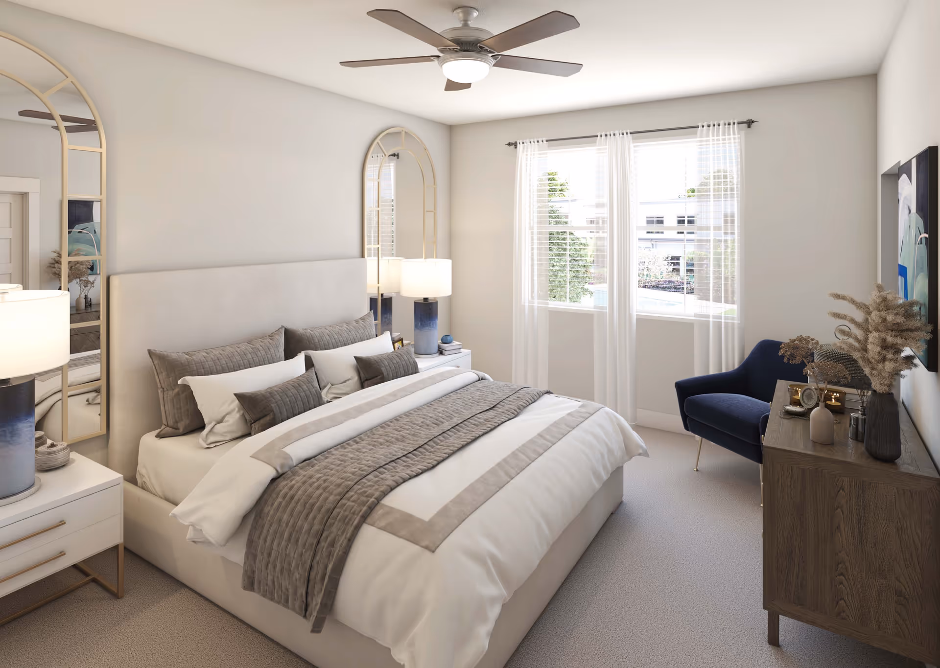 A bright and cozy bedroom with a large bed featuring white and beige bedding and multiple pillows. There are two tall arched mirrors behind the nightstands on either side of the bed, each with a lamp. A ceiling fan with light is mounted on the ceiling. A window with sheer white curtains lets in natural light. A wooden dresser with decorative items and a blue armchair are positioned against the wall.