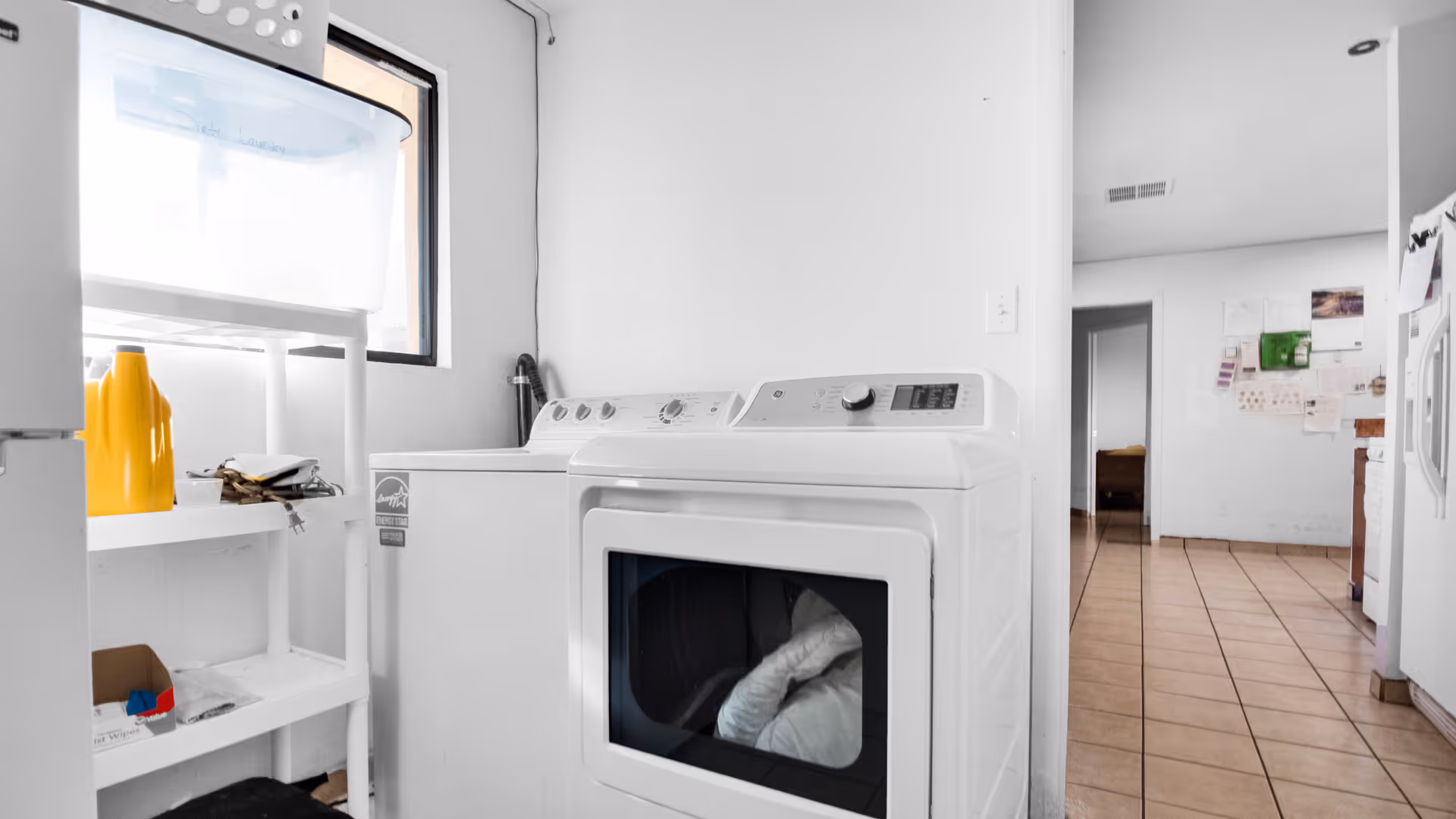 Laundry room with a white washing machine and dryer side by side, a white shelving unit holding a yellow container and other items, a window letting in natural light, and a view into an adjacent room with tiled floor and white walls.