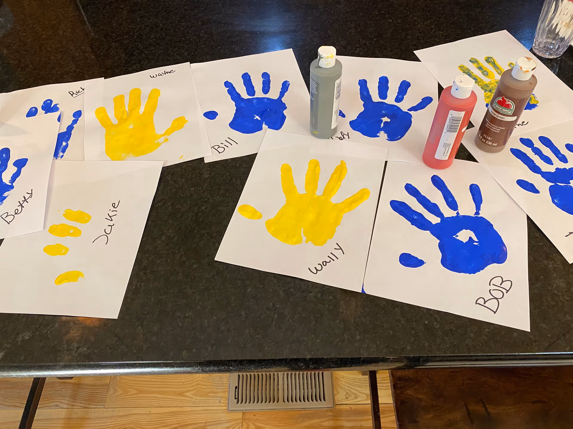 Yellow and blue painted handprints on sheets of paper with bottles of paint on a dark tabletop.