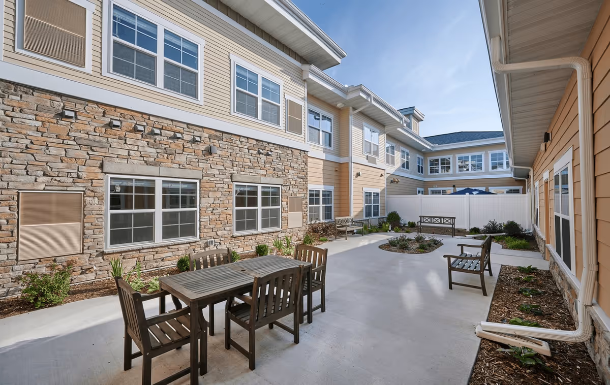 Outdoor courtyard area at Stonebay Senior Living featuring wooden tables and chairs on a concrete patio surrounded by a two-story building with stone and beige siding, multiple windows, and some landscaping with plants and shrubs.