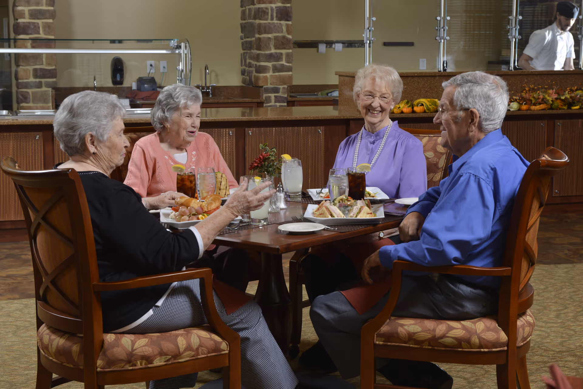 Four elderly people sitting around a dining table in a retirement community, enjoying a meal and drinks together. The setting includes wooden chairs with patterned cushions and a counter with a brick pillar and a staff member in the background.