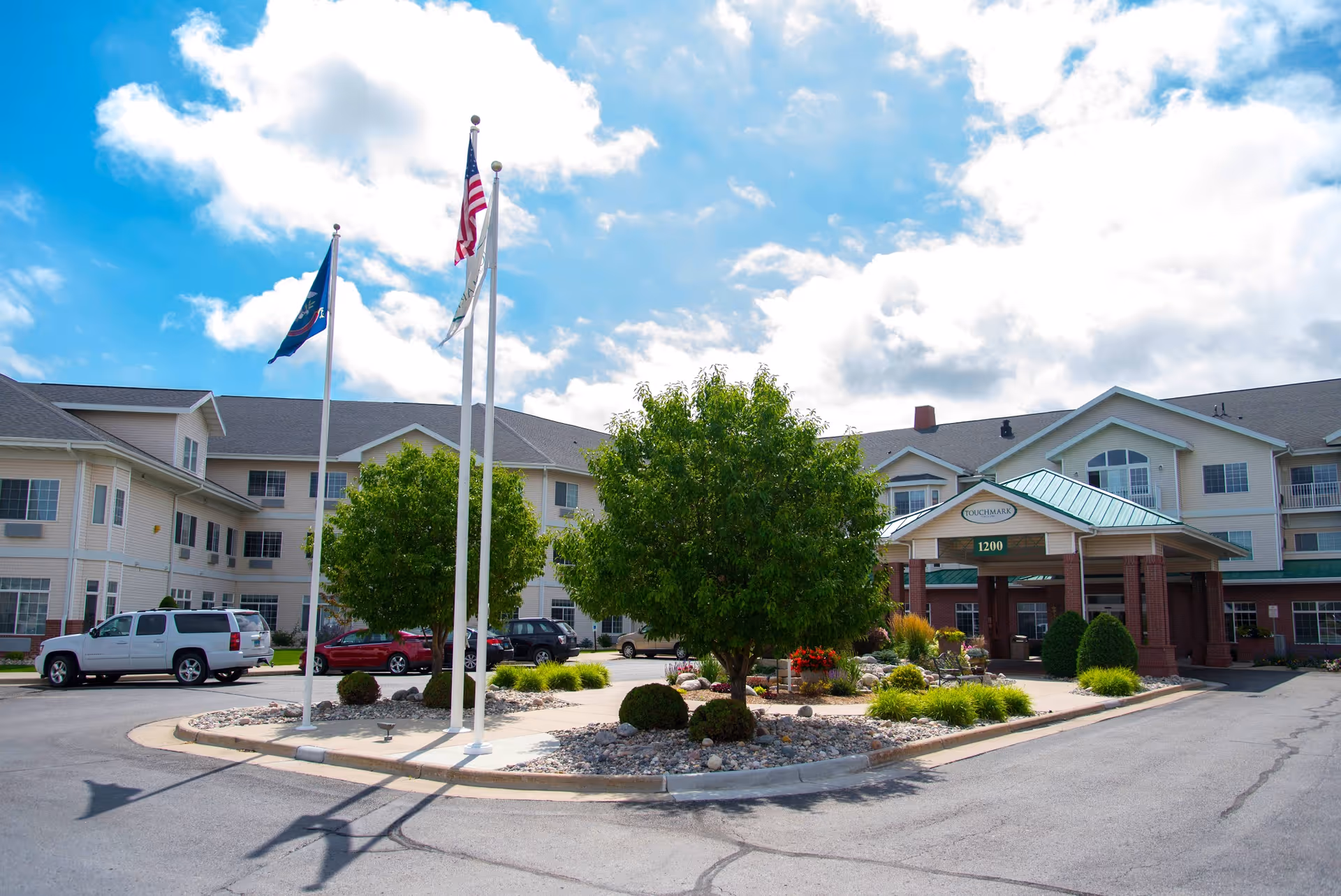 Exterior view of Touchmark at Harwood Groves senior living facility showing a large building with multiple windows, a covered entrance with the facility name and address 1200, a landscaped roundabout with trees and shrubs, and three flagpoles with flags against a partly cloudy blue sky.