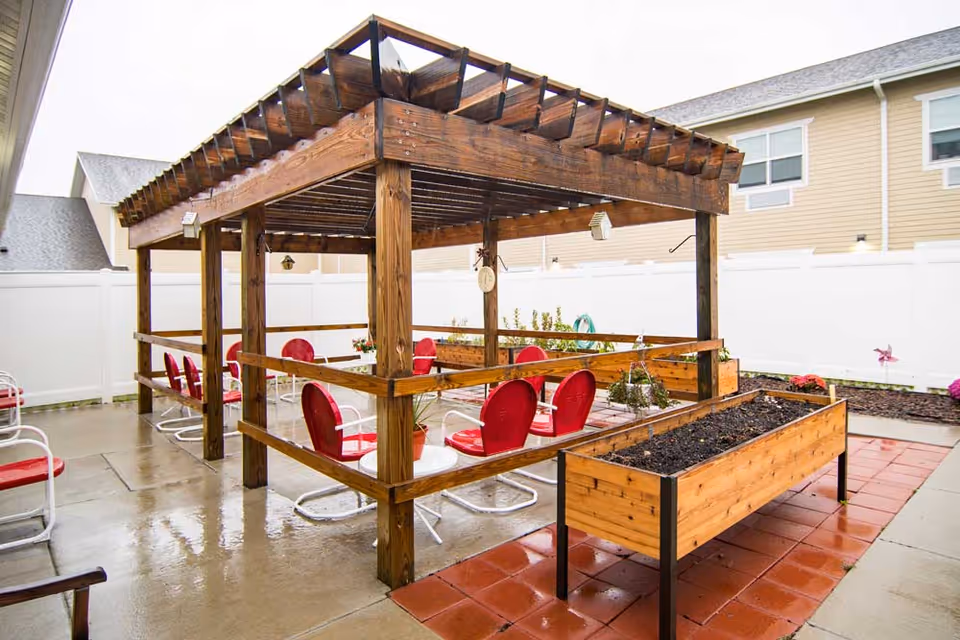 Outdoor patio area with a wooden pergola structure, red and white metal chairs arranged around small tables, and raised garden beds with soil. The area is surrounded by a white fence and residential buildings are visible in the background.