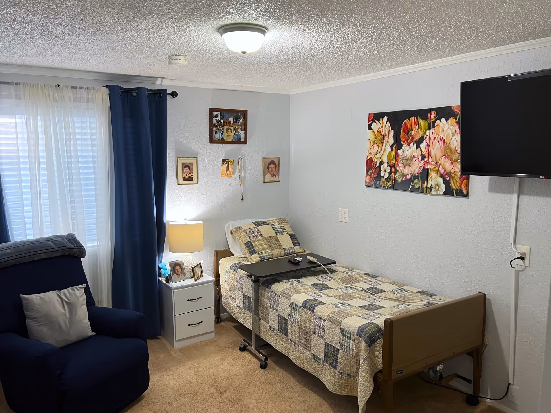 A cozy bedroom in a senior care facility featuring a single bed with a patchwork quilt, a small bedside table with a lamp and framed photos, a navy blue armchair with a gray pillow, blue curtains over a window with white blinds, a wall-mounted flat-screen TV, and floral artwork on the wall.