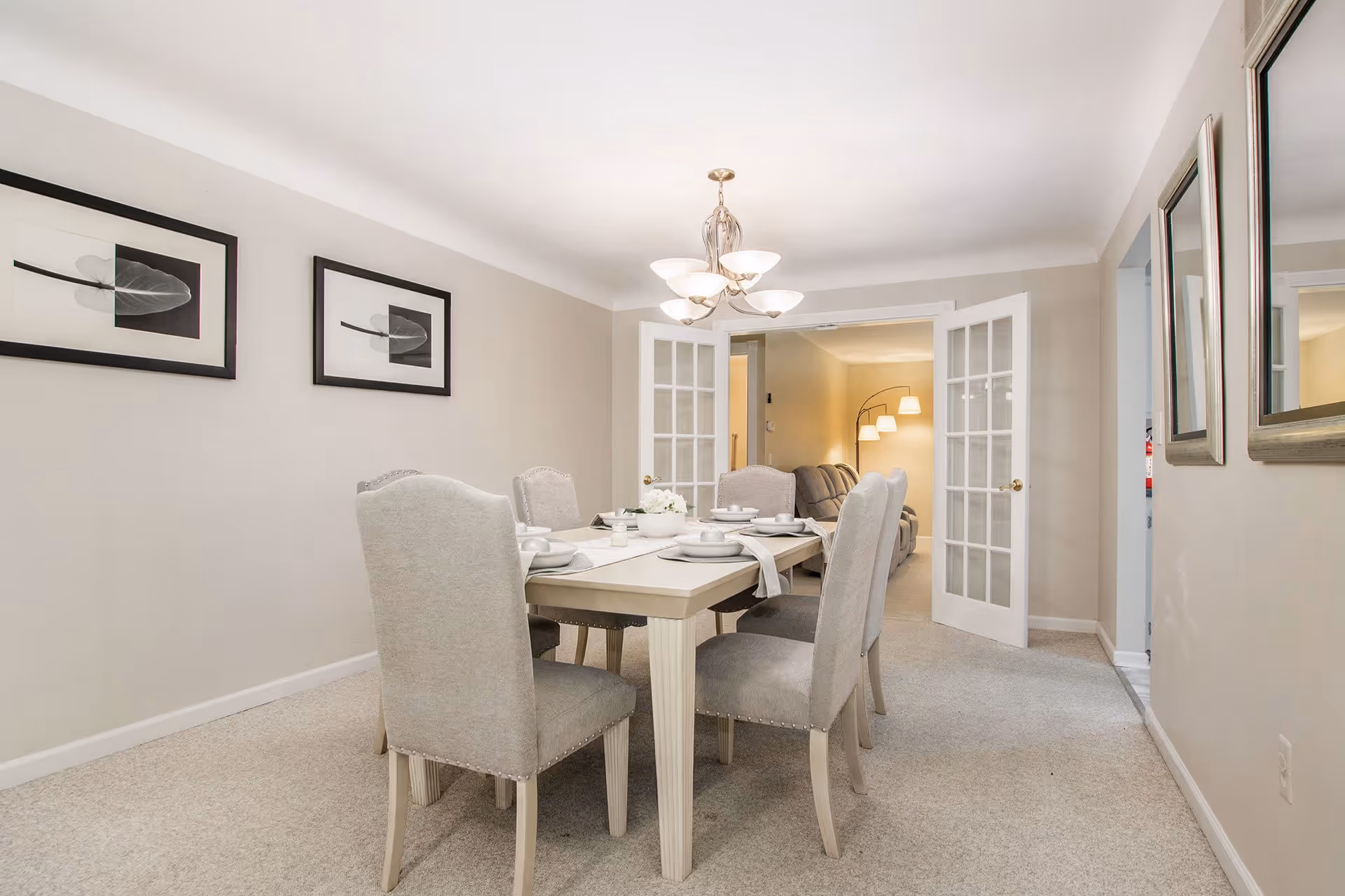 A dining room with a rectangular table set for six people with white plates and napkins. The room has beige walls, carpeted floor, two framed leaf prints on the left wall, and a chandelier hanging above the table. French doors open into a living room area with a sofa and a floor lamp with multiple lights.