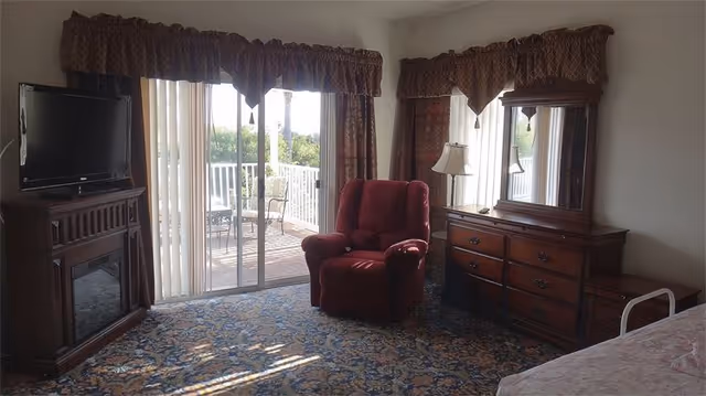 A cozy living room area with a red recliner chair, a wooden dresser with a mirror, a TV on a wooden stand with an electric fireplace, and sliding glass doors leading to an outdoor patio with chairs and a table. The room has patterned curtains and a floral carpet.