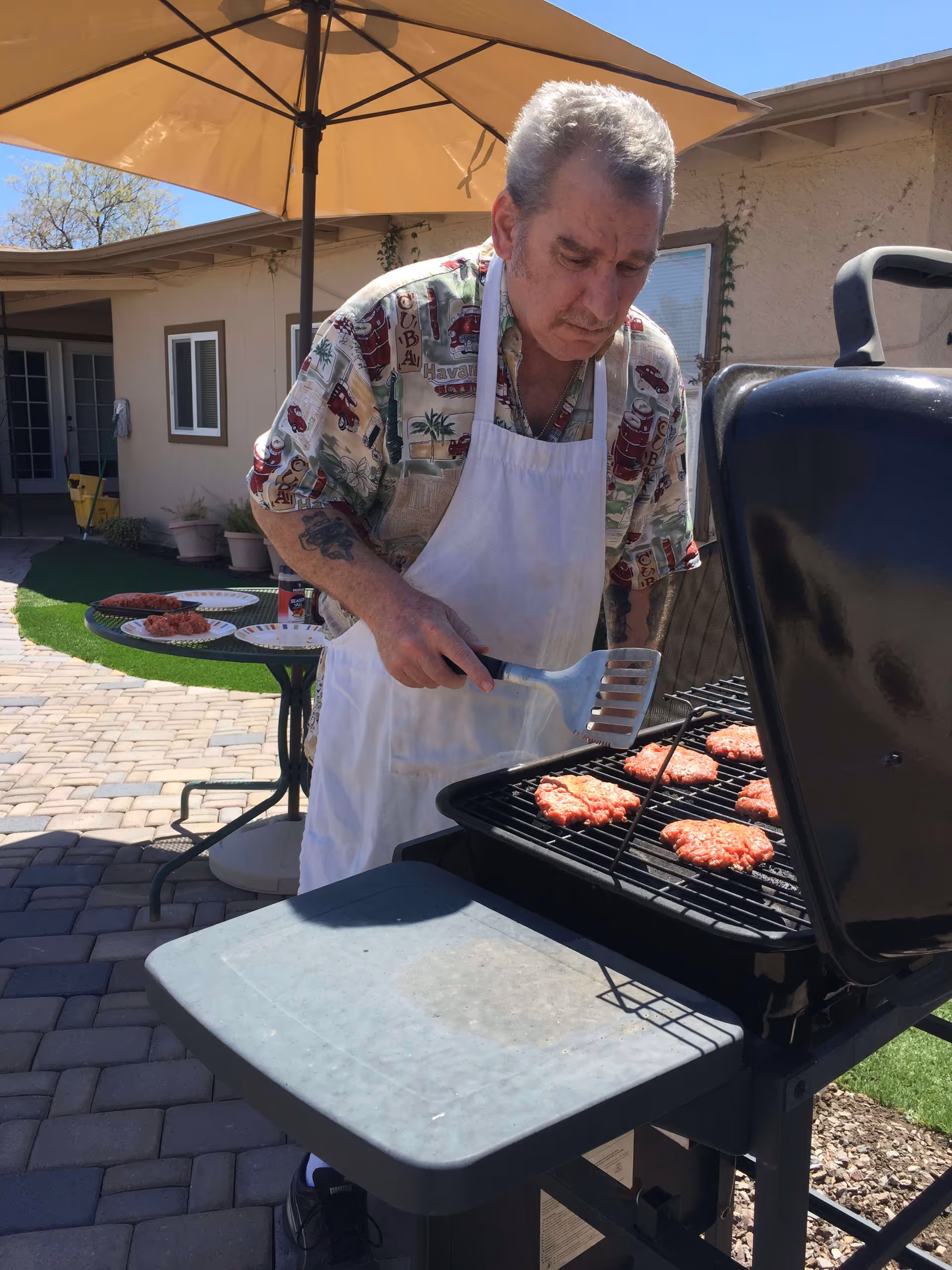 A man wearing a white apron and a patterned shirt is grilling several hamburger patties on a black outdoor grill under a large beige patio umbrella. Behind him, there is a building with windows and a door, and a round table with plates of raw hamburger patties.