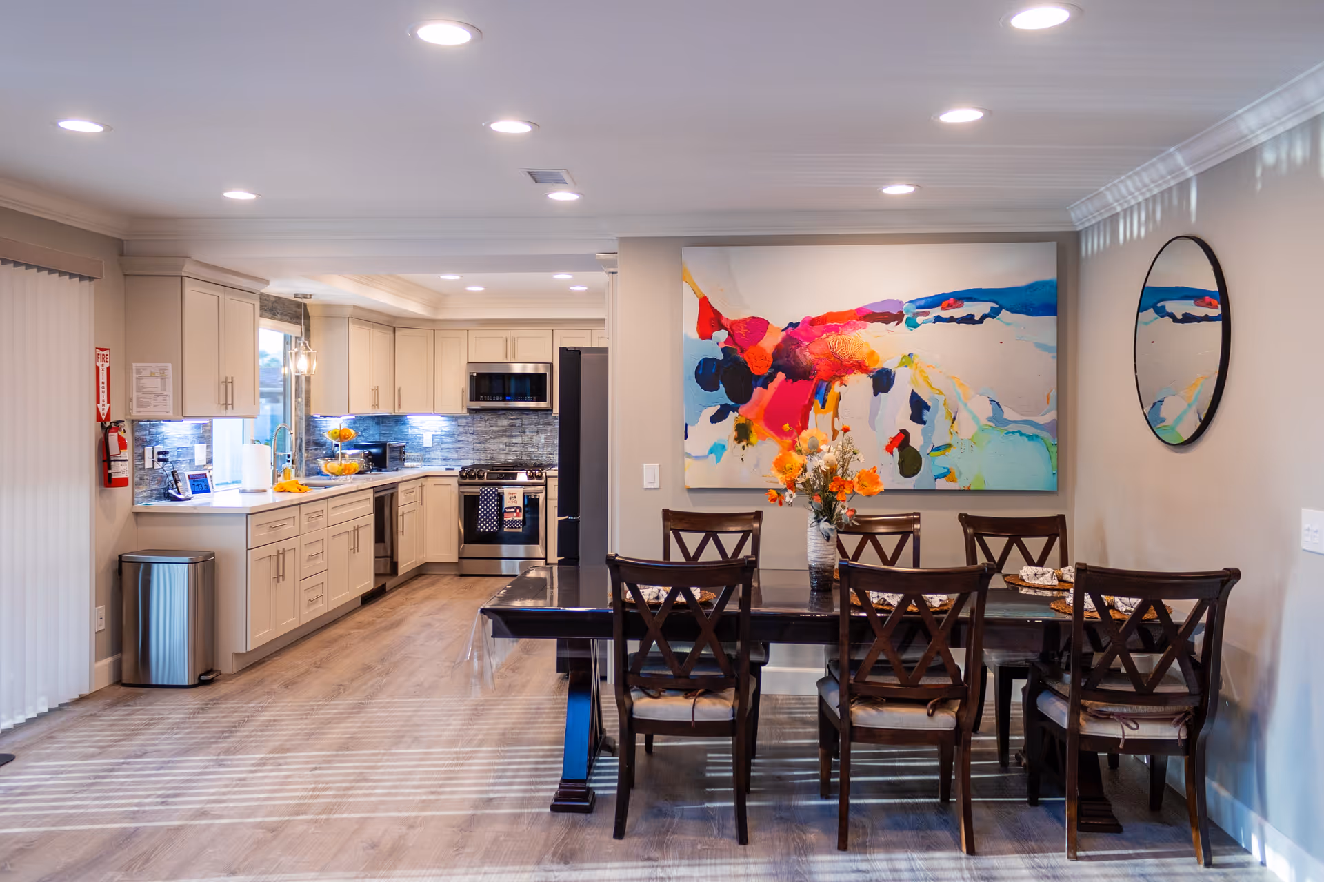 A bright and modern open kitchen and dining area in a senior living facility. The kitchen features light-colored cabinets, stainless steel appliances including a microwave, stove, and refrigerator, and a tiled backsplash. The dining area has a dark wooden table with six matching chairs, a vase with flowers on the table, a large colorful abstract painting on the wall, and a round mirror. The room has recessed lighting and wood flooring.