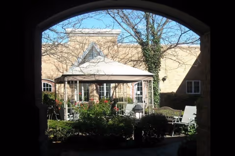 View through an arched walkway into a sunny courtyard with a gazebo, patio chairs, shrubs, and a brick building.