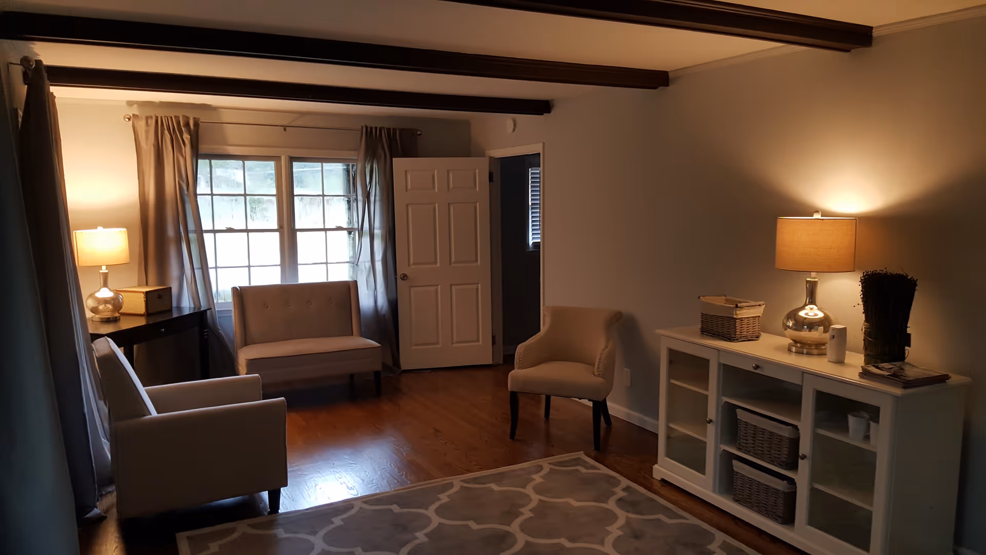 A cozy living room with wooden floors, a patterned area rug, and three beige upholstered chairs. There is a white cabinet with glass doors on the right side, topped with a lamp, a basket, and decorative items. A window with curtains and a door are visible in the background, with another lamp on a small table near the window.