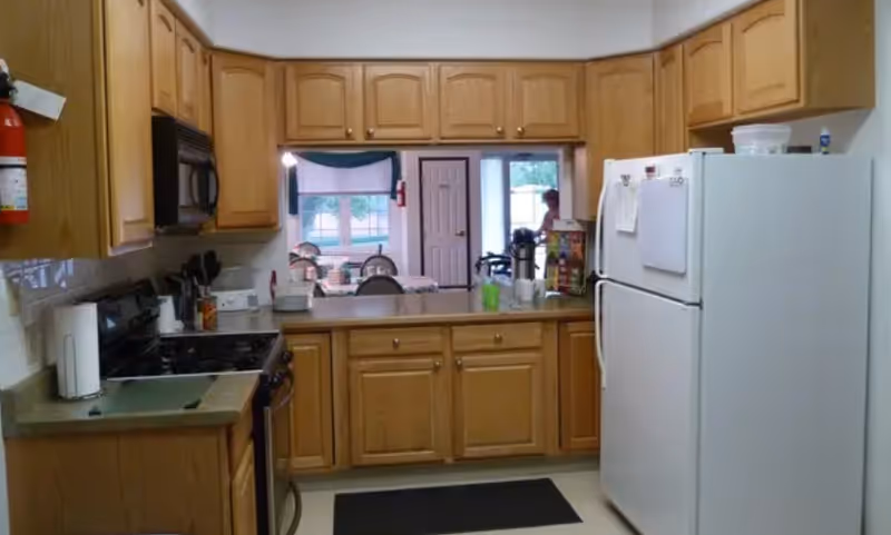 A kitchen with wooden cabinets, a white refrigerator, a stove with a microwave above it, and a countertop with various items. In the background, there is a dining area with a table and chairs, and a person is visible near the doorway.