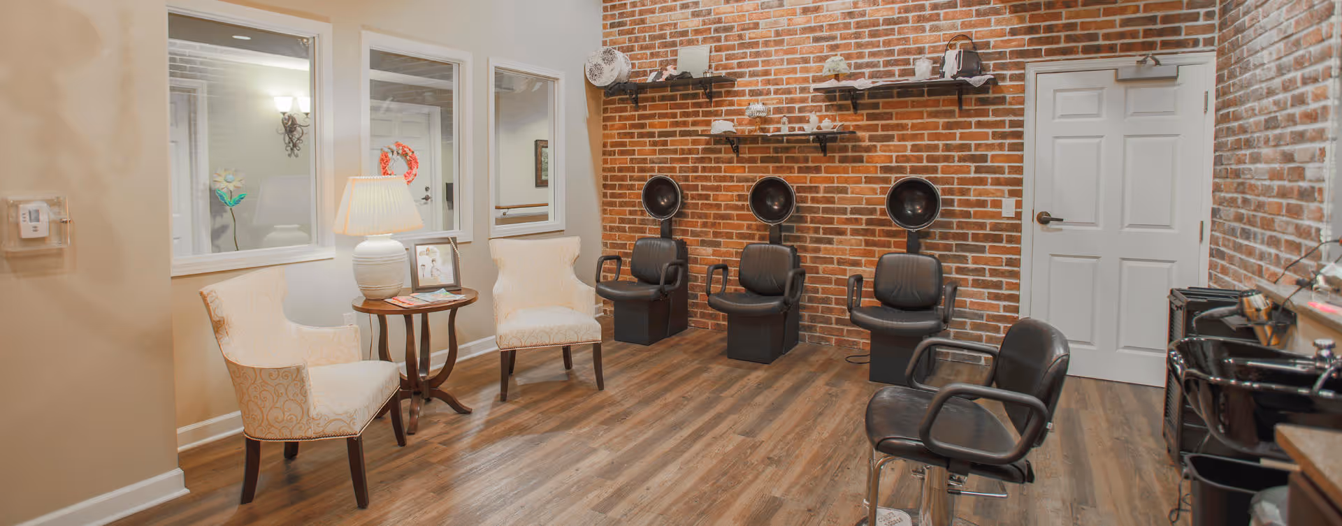 Interior of a salon area with three black hair dryer chairs against a brick wall, two beige upholstered chairs with a small round table and lamp between them, and a black salon chair in the foreground. The room has wood flooring and a white door on the right side.