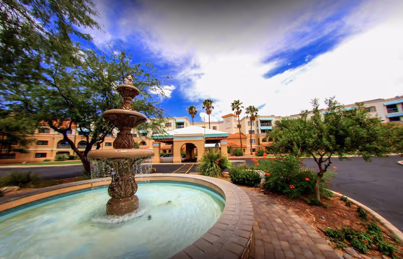 Front entrance of a multi-story senior living building with a tiered fountain, landscaped driveway, and palm trees under a blue sky.