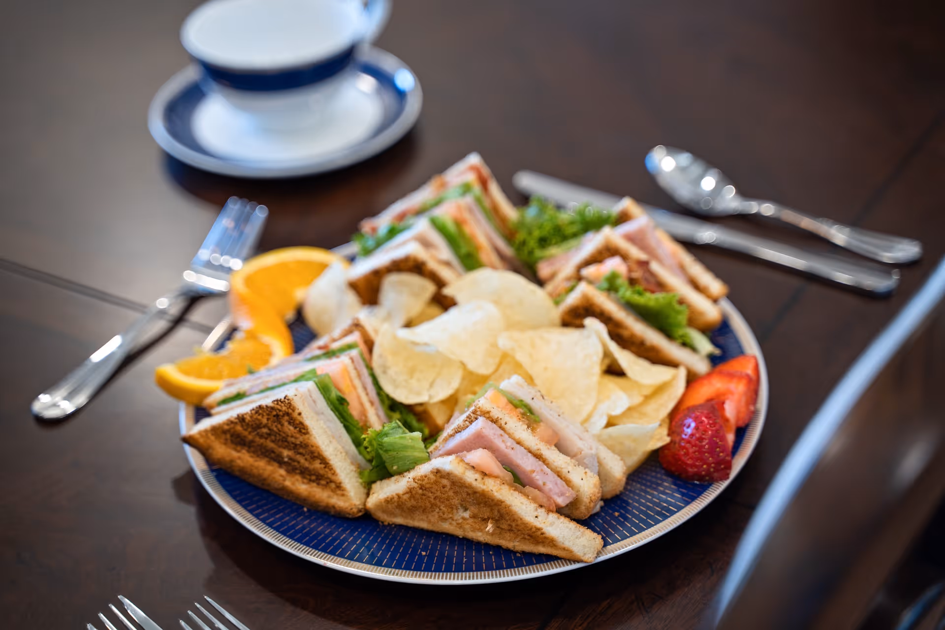 A plate with several club sandwich quarters filled with lettuce, tomato, and deli meat, accompanied by potato chips, orange slices, and a strawberry, placed on a dark wooden table with a cup and saucer, fork, and spoon nearby.
