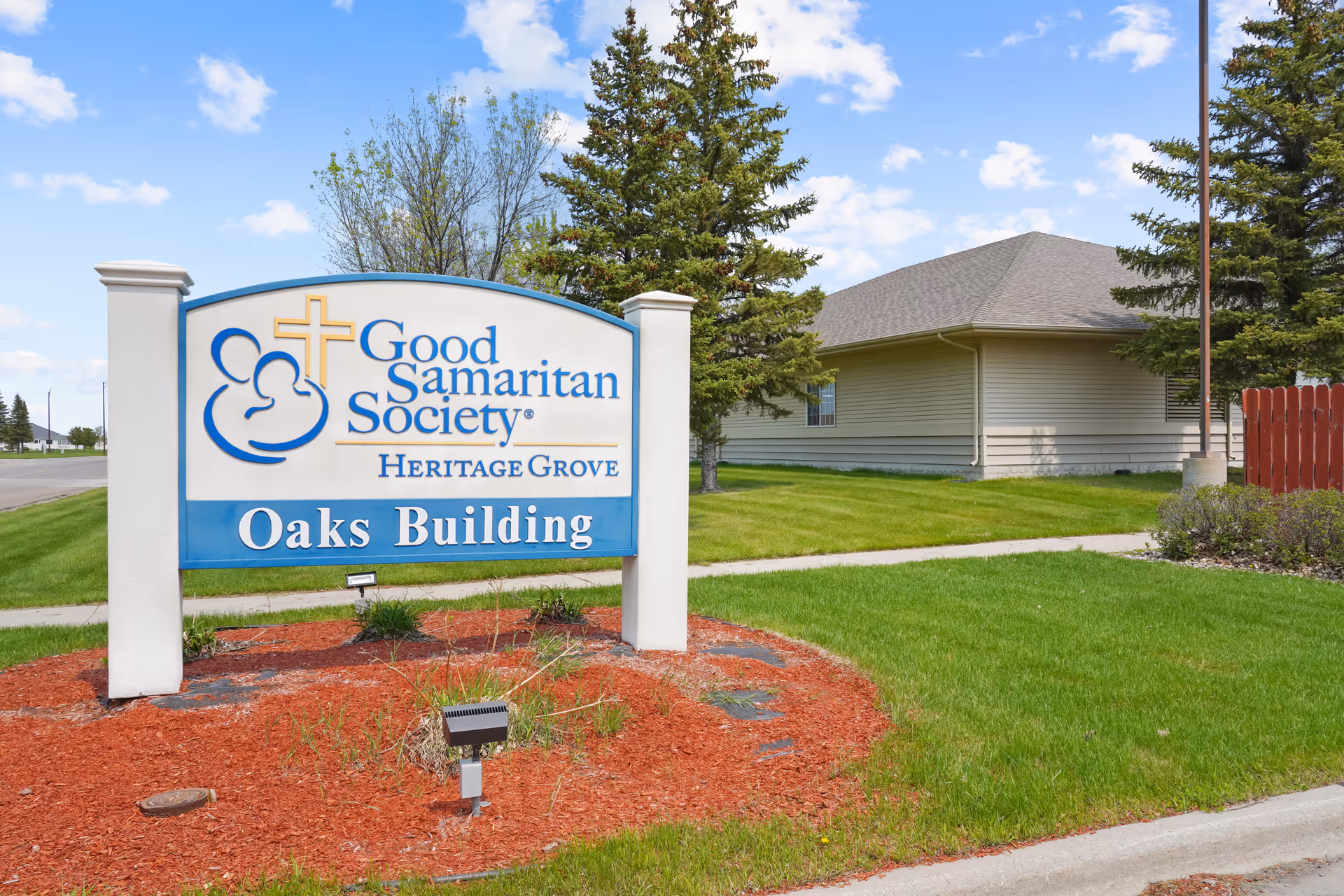 A Good Samaritan Society - Heritage Grove 'Oaks Building' sign on a landscaped lawn in front of a single-story building and trees.
