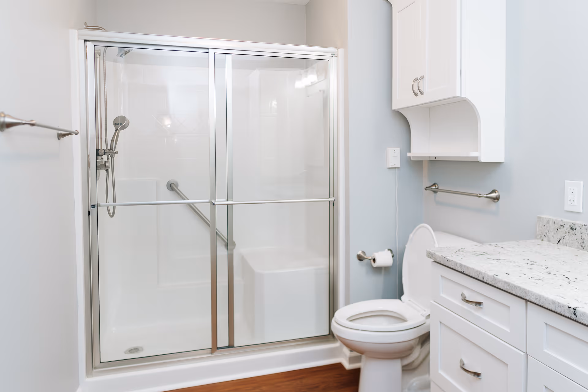 A clean and modern bathroom featuring a glass-enclosed shower with a handheld showerhead and built-in seat, a white toilet with the lid open, a granite countertop with white cabinetry, and a wall-mounted cabinet above the toilet. The walls are painted light gray and the floor has a wood-like finish.