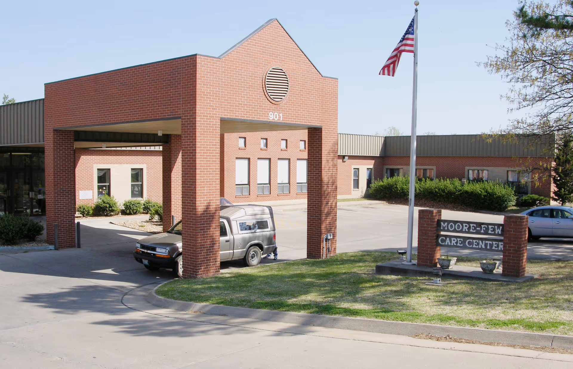 Exterior view of Moore-Few Care Center showing a brick entrance structure with the number 901, a flagpole with an American flag, a sign with the facility name, and parked vehicles in the driveway.