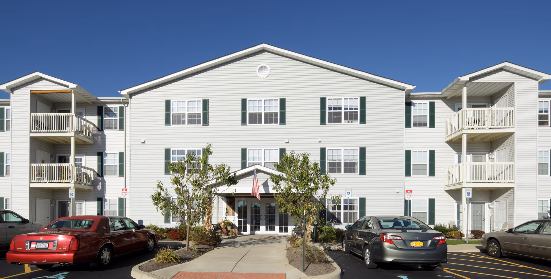 Exterior front view of South Pointe Senior Apartments, a three-story white building with green shutters and balconies on each side. There are several parked cars in front, a walkway leading to the entrance, small trees, and an American flag above the main door under a clear blue sky.