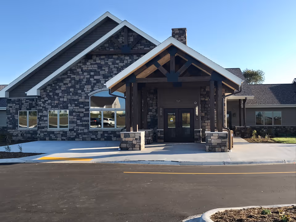 Front exterior view of a building with stone and wood accents, featuring a covered entrance with double doors and large windows reflecting the surrounding area under a clear blue sky.