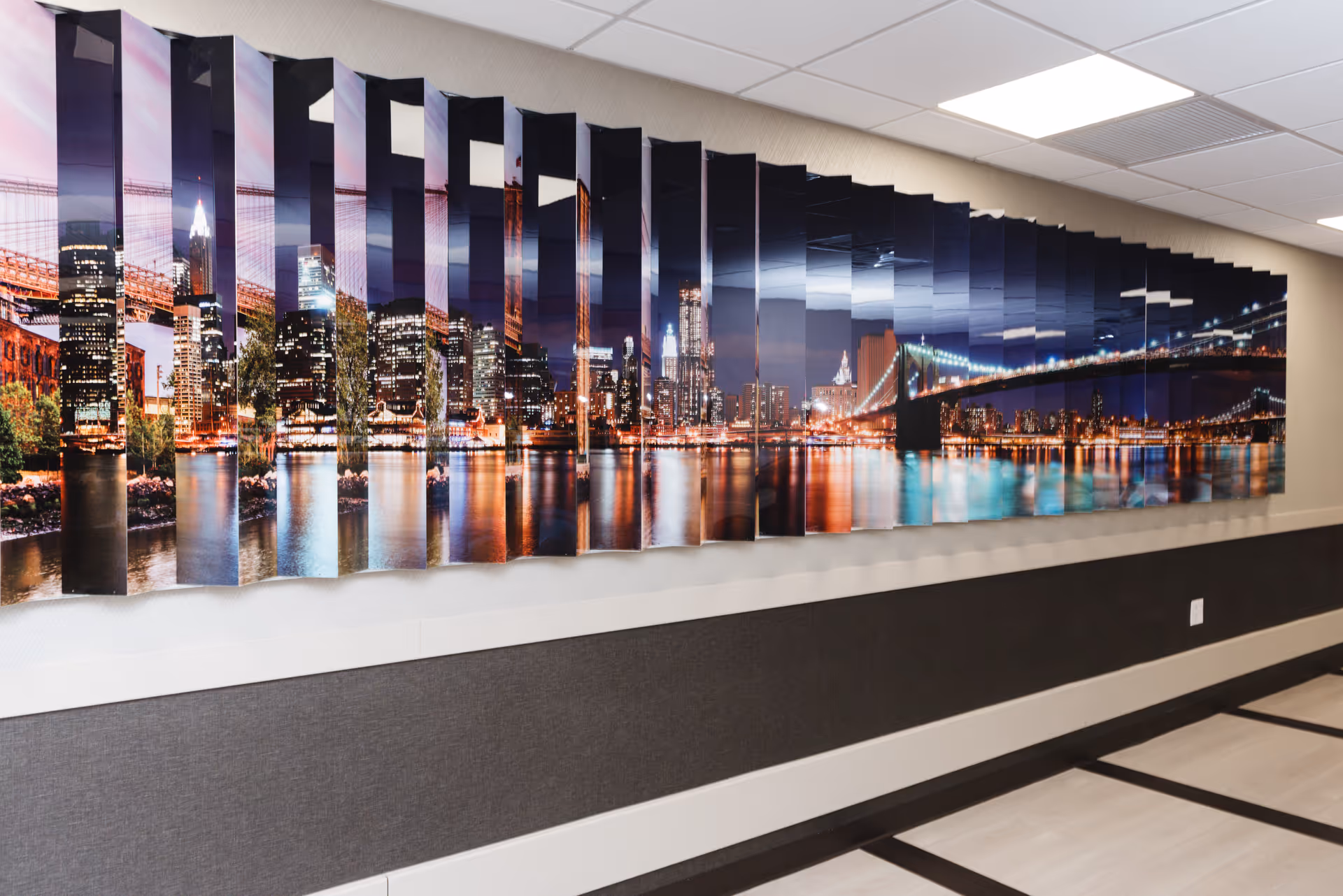 A long, modern hallway wall decorated with a large, segmented panoramic artwork depicting a nighttime city skyline with illuminated buildings and a bridge reflected in the water. The wall has a two-tone design with a dark lower section and a light upper section, and the ceiling features recessed lighting panels.