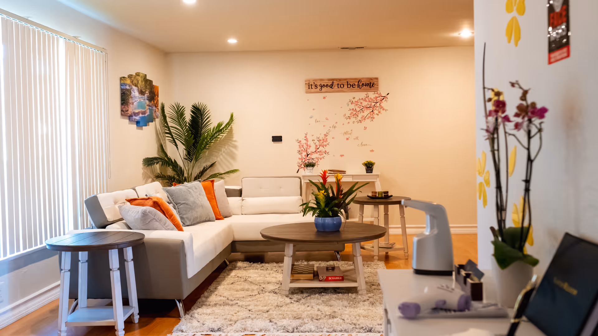 A cozy living room with a white and gray sectional sofa adorned with orange and gray cushions. There is a round wooden coffee table with a potted plant on it, a side table, and a white rug on a wooden floor. The wall features a decorative sign that reads 'it's good to be home' along with floral wall decals. A large green plant is placed in the corner near vertical blinds covering a window. Additional decor includes a small table with books and plants, and a white cabinet with a few items on top.