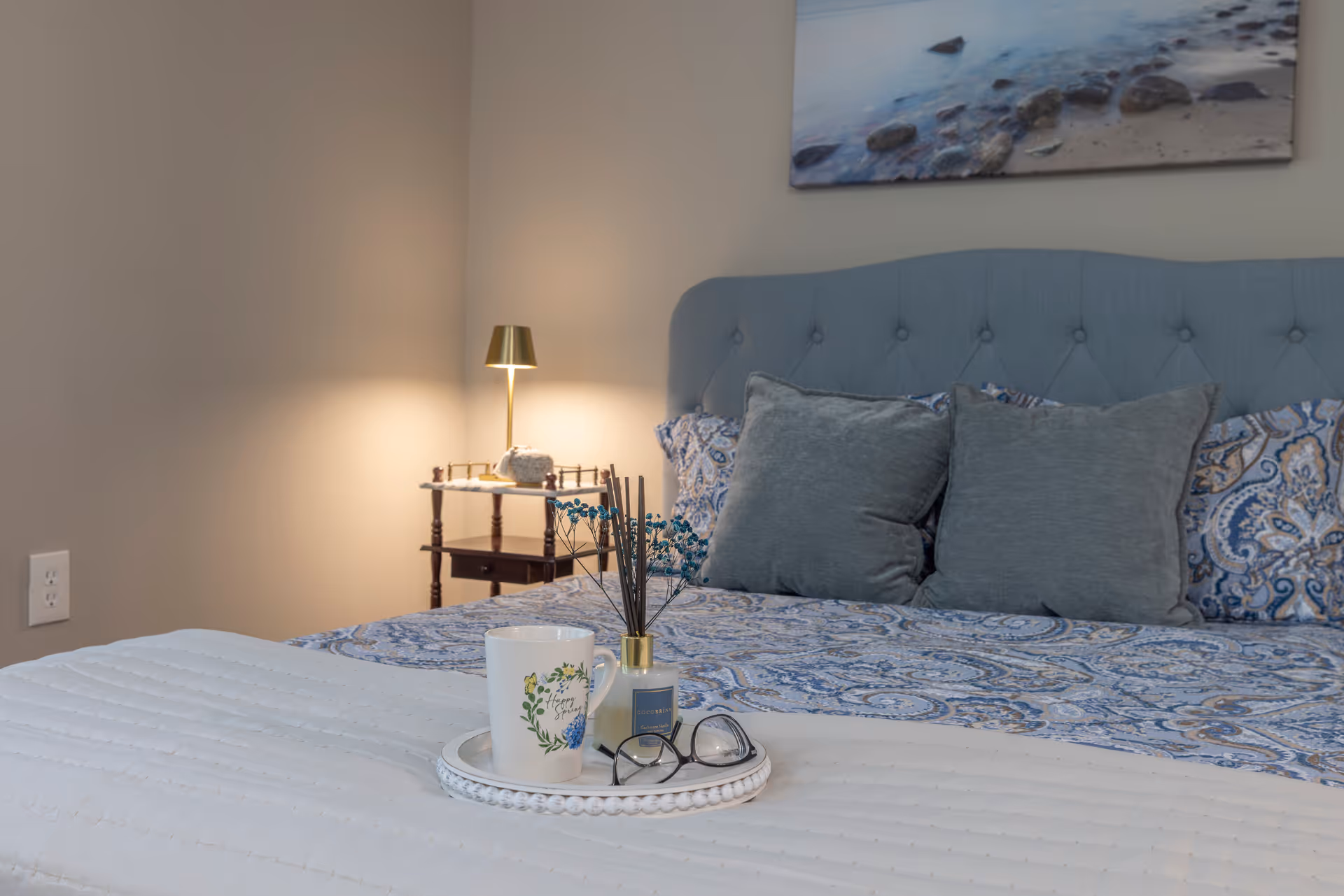 A cozy bedroom featuring a bed with a blue patterned bedspread and two gray pillows. A small round tray on the bed holds a white mug, a reed diffuser, and a pair of eyeglasses. In the background, there is a wooden nightstand with a gold-colored lamp and a decorative item. A framed picture of a rocky shoreline hangs on the wall above the bed.