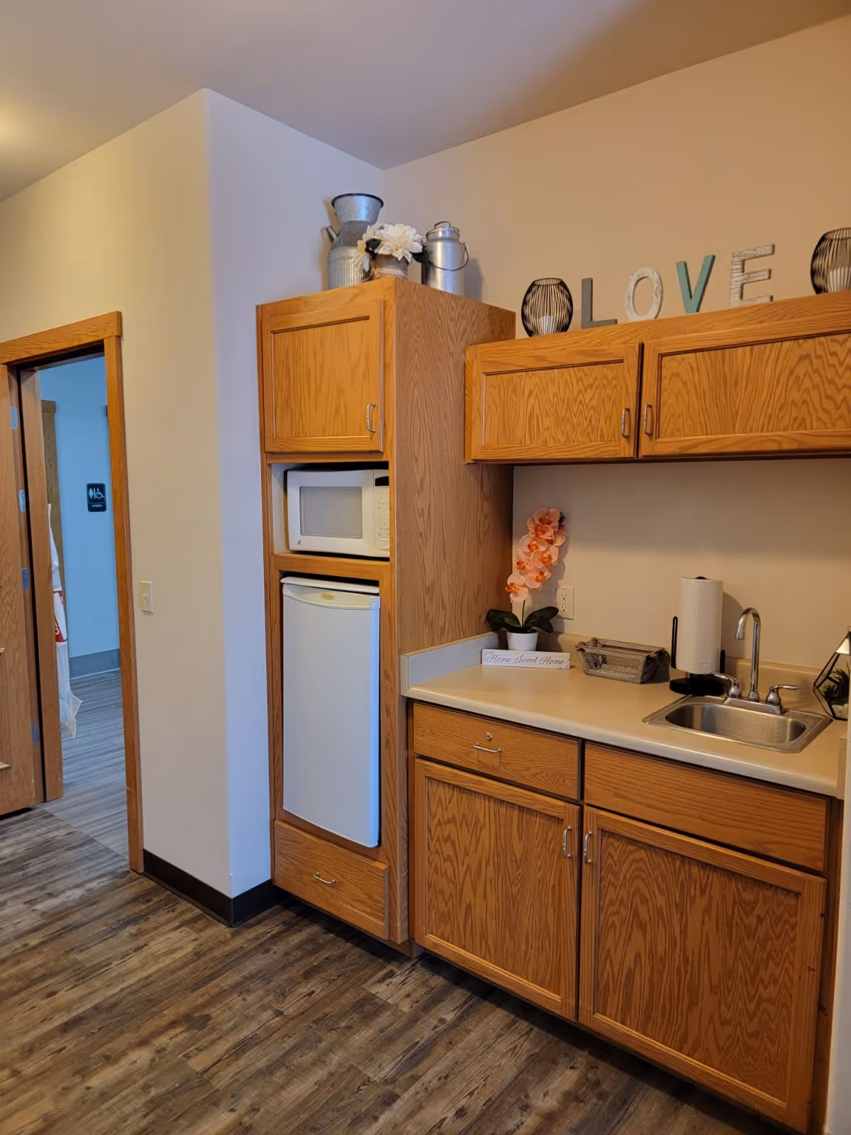 A small kitchen area with wooden cabinets, a white microwave, and a mini refrigerator. The countertop has a sink, a paper towel holder, a small basket, and a decorative pink orchid plant. Above the cabinets, there are decorative items including metal containers and letters spelling 'LOVE'. The floor is wood-patterned, and there is a doorway leading to another room.