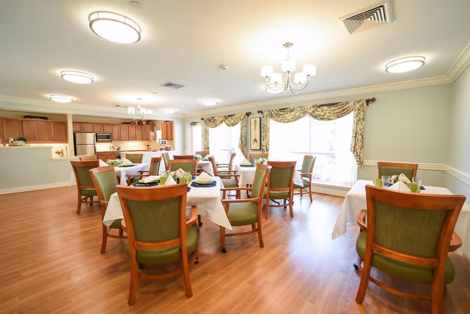 A bright dining room in a senior living facility with several tables covered in white tablecloths, each set with green napkins and tableware. The room features wooden chairs with green upholstery, large windows with floral curtains, and a kitchen area with wooden cabinets and stainless steel appliances in the background.