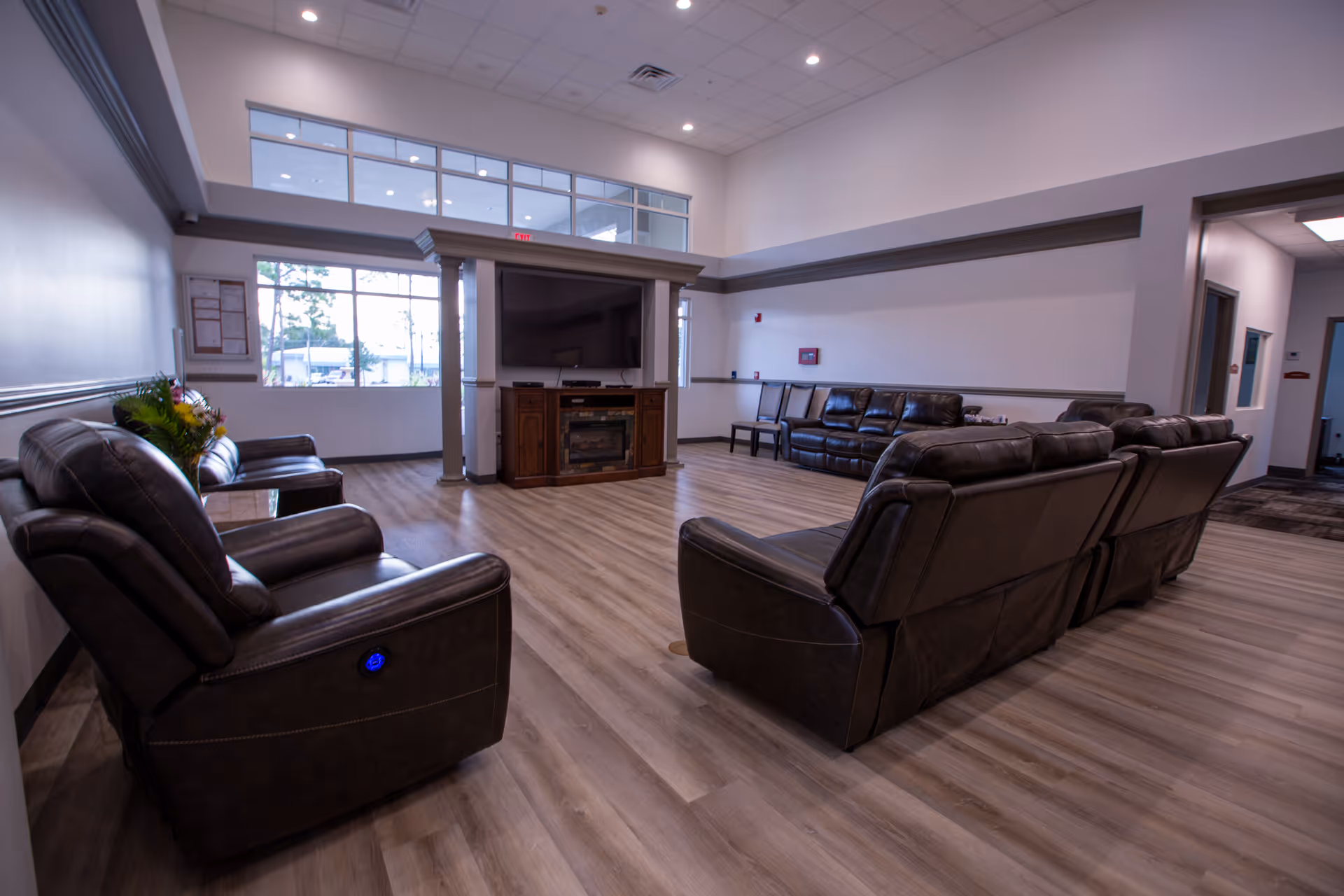 A spacious living room area with multiple dark brown leather recliners and sofas arranged around a large flat-screen TV mounted on a wooden entertainment center. The room has light wood flooring, white walls, and large windows letting in natural light. There are also some chairs lined up against the far wall and a hallway visible to the right.