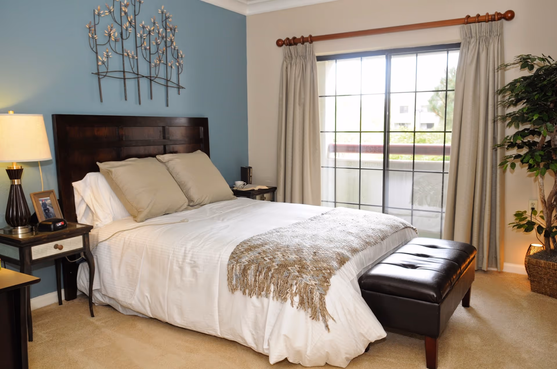 A cozy bedroom with a large bed featuring white bedding and beige pillows. There is a dark wooden headboard and a matching bench at the foot of the bed. On the left side, there is a nightstand with a lamp, a framed photo, and an alarm clock. The wall behind the bed is painted blue and decorated with a metal wall art piece. To the right, there is a large window with beige curtains and a potted plant in the corner.