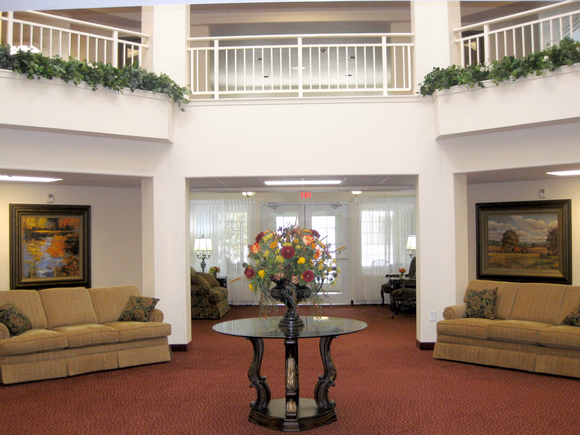 A spacious and well-lit common area with two beige sofas facing each other, a round glass-top table with a large floral arrangement in the center, framed landscape paintings on the walls, and a second-floor balcony with white railings and green plants.