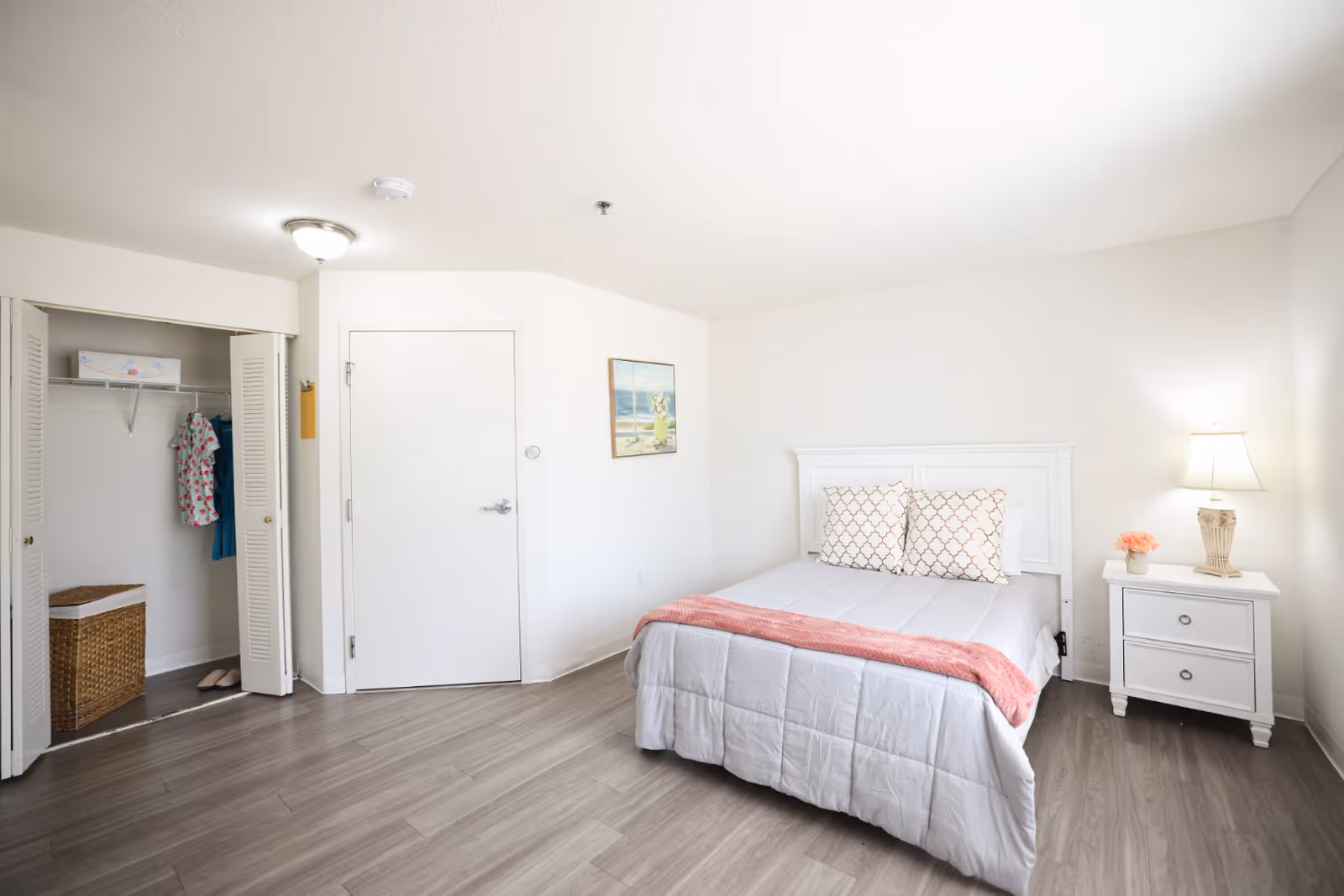 A bright and clean bedroom with a white bed featuring two patterned pillows and a pink throw blanket. To the right of the bed is a white nightstand with a lamp and a small vase of flowers. On the left side of the room, there is an open closet with a wicker basket and a couple of hanging clothes. The walls are white, and there is a framed painting of a beach scene hanging above the bed. The floor is covered with light wood laminate.