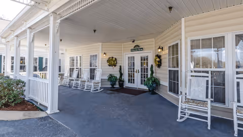 Covered porch area of a senior living facility with white rocking chairs lined up along the wall, potted plants, and a double glass door entrance with a sign above it reading 'The Bungalows'.