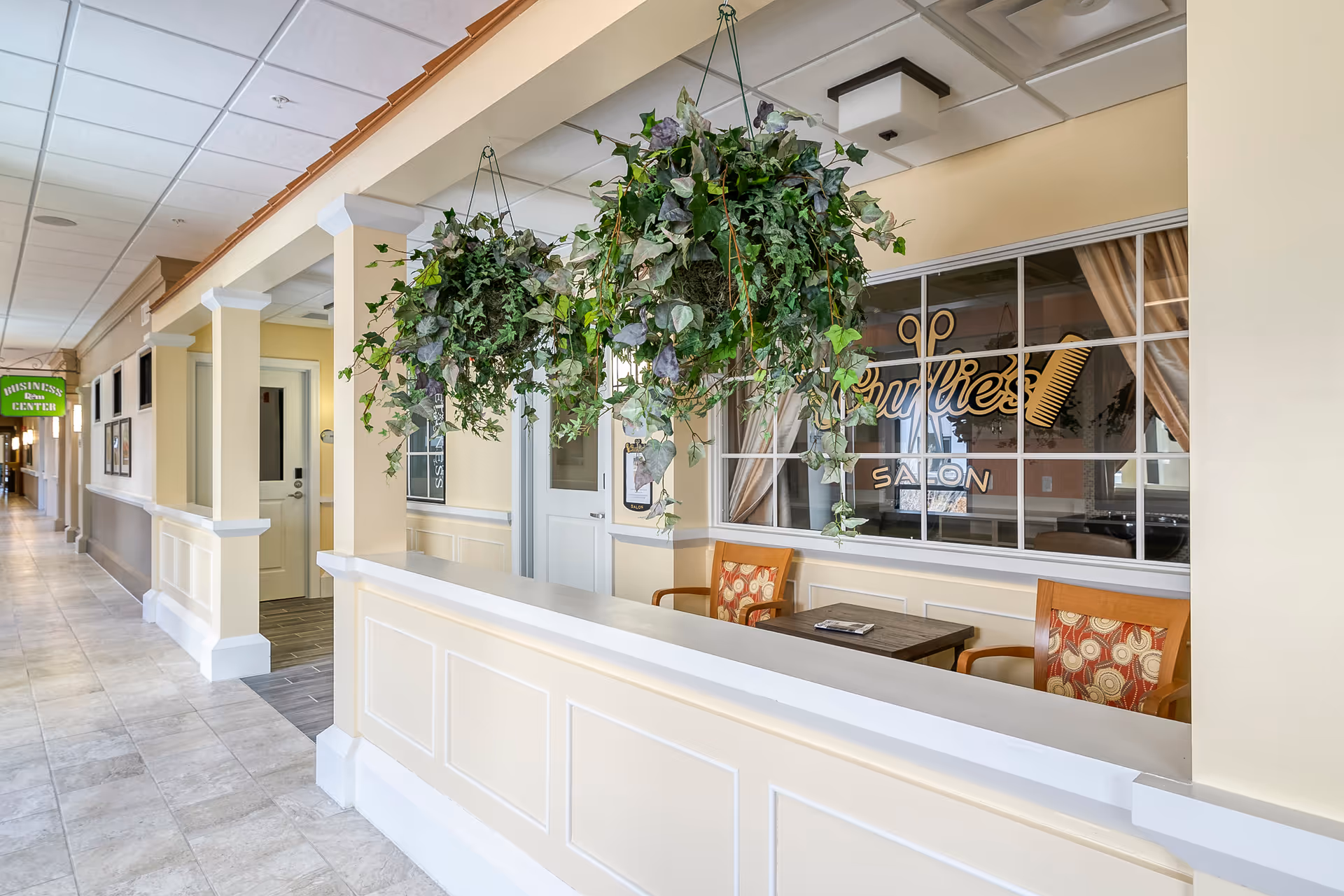 Interior hallway of Willowbrook Hills facility featuring a reception or waiting area with hanging green plants, two chairs with patterned cushions, and a window displaying the sign 'Sullie's Salon'. The hallway extends to the left with a sign indicating a business center.
