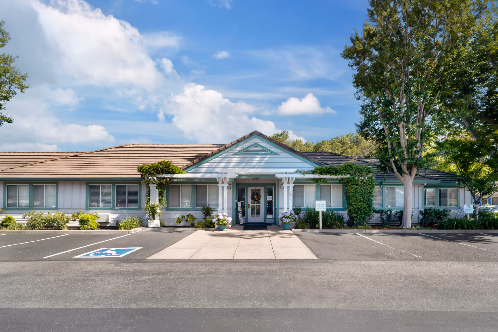 Single-story senior living building facade with a central entrance, landscaping, and a parking lot with a marked handicap space.