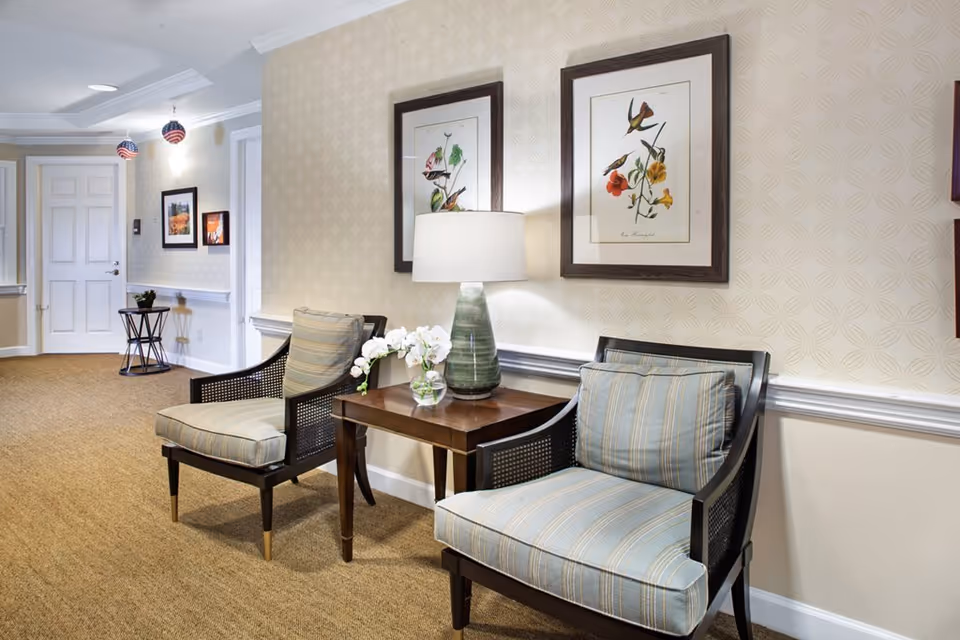 Two upholstered chairs flanking a wooden side table with a lamp and flowers in a well-lit senior living facility hallway.