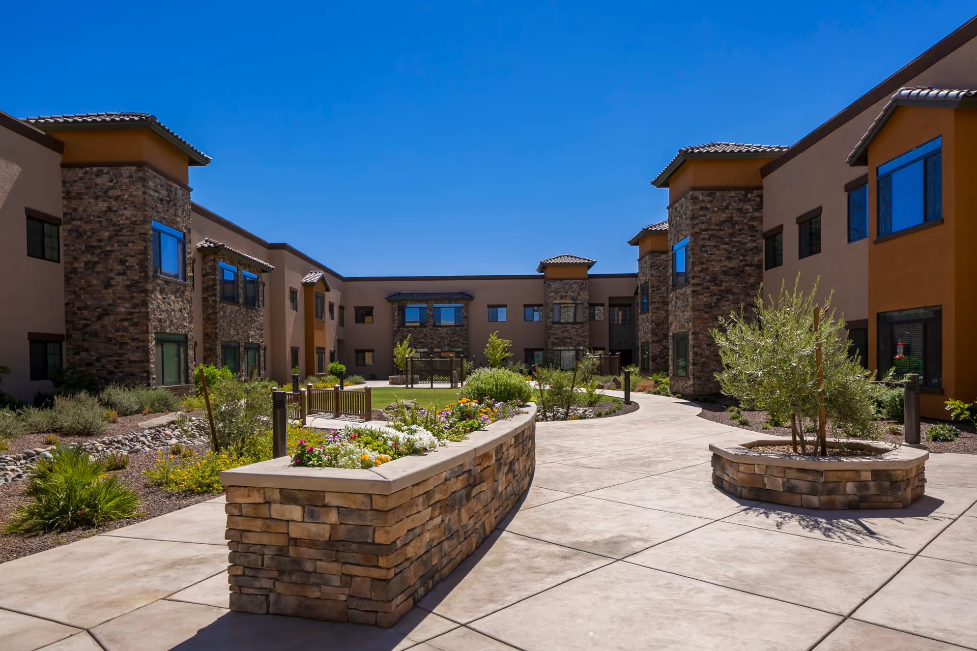 Outdoor courtyard area of Orchard Pointe at Arrowhead featuring a paved walkway, raised stone flower beds with colorful flowers and small trees, surrounded by a two-story building with stone and stucco exterior under a clear blue sky.