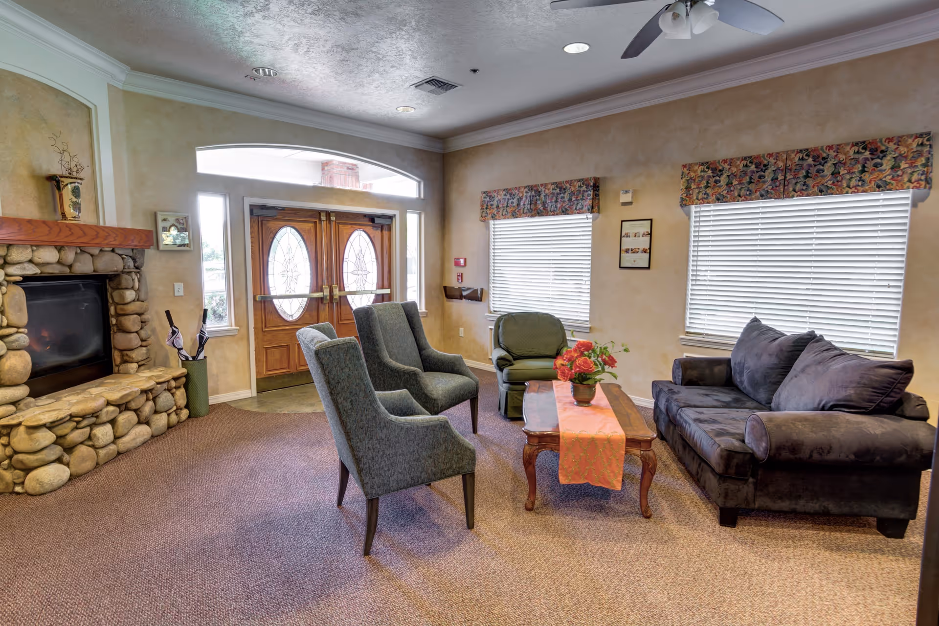A cozy senior living facility common area with a stone fireplace on the left, two gray armchairs, a green armchair, and a dark gray sofa arranged around a wooden coffee table with a floral centerpiece. The room has beige walls, two windows with floral valances, and double wooden doors with oval glass panels letting in natural light.