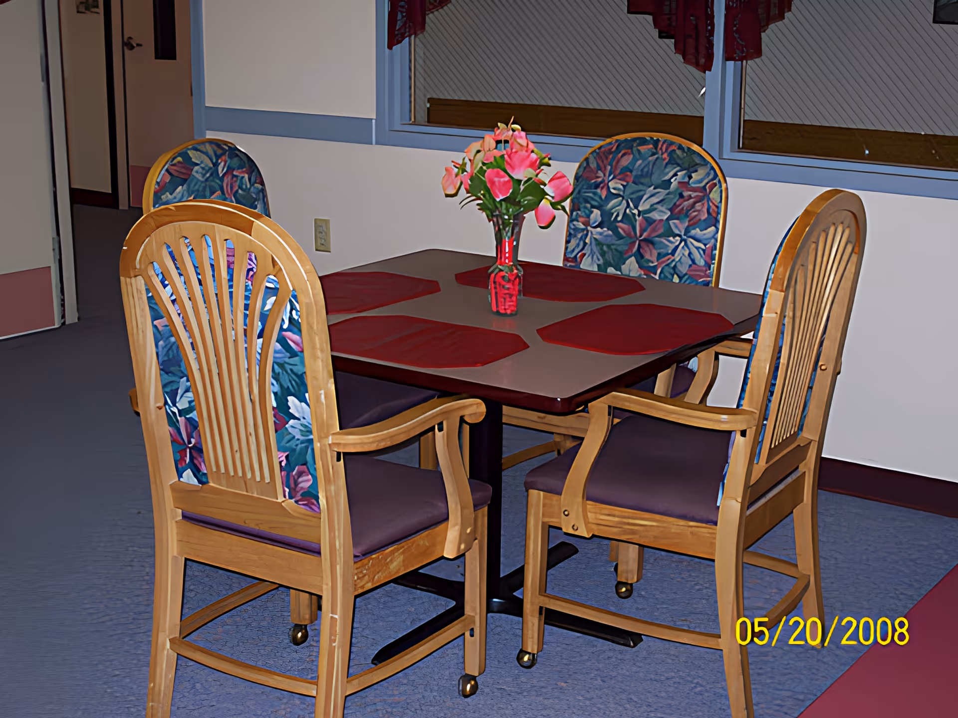 A small dining table set with four wooden chairs and a vase of pink flowers in a communal room.