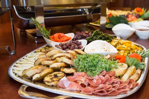 A large silver platter filled with an assortment of grilled vegetables, sliced meats, olives, and fresh greens arranged attractively on a buffet table.