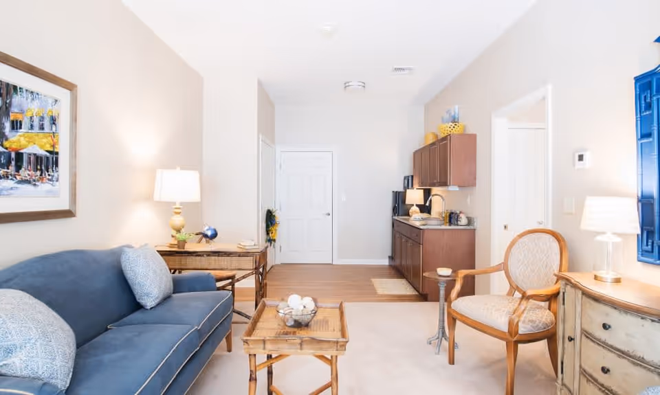 A bright and cozy living room area with a blue sofa adorned with patterned cushions on the left, a wooden coffee table with decorative items in the center, and a wooden armchair with a cushioned seat on the right. There is a side table with a lamp next to the sofa and another lamp on a small dresser on the right. The room has light-colored walls, a framed painting above the sofa, and a small kitchenette with wooden cabinets and a countertop in the background.