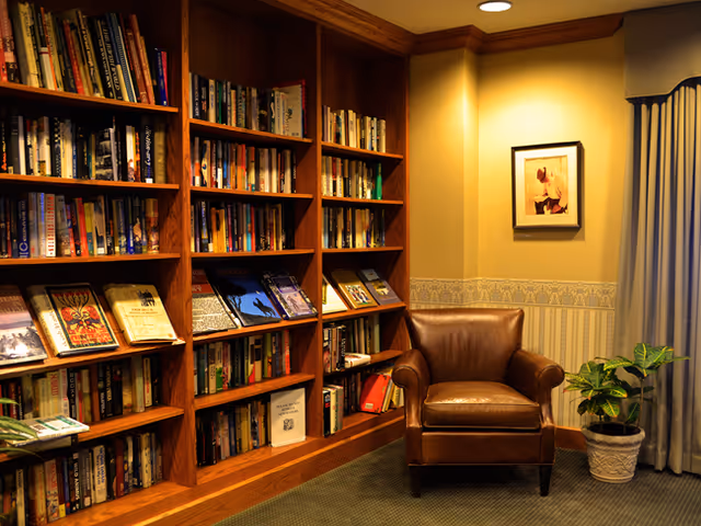 Cozy reading nook with wooden bookshelves filled with books, a leather armchair, potted plant, and framed artwork on the wall.