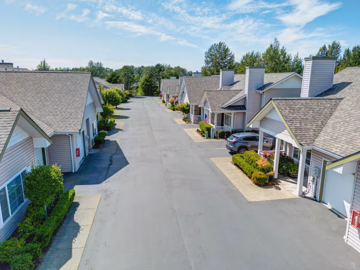 View of a residential street within a senior living community showing single-story houses with garages, small front porches, and well-maintained landscaping under a partly cloudy sky.