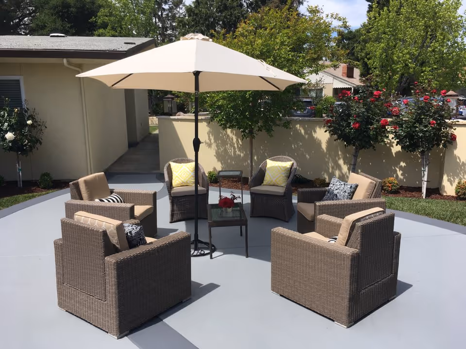 Outdoor patio with wicker chairs arranged in a circle under a beige umbrella next to a low wall and rose bushes.