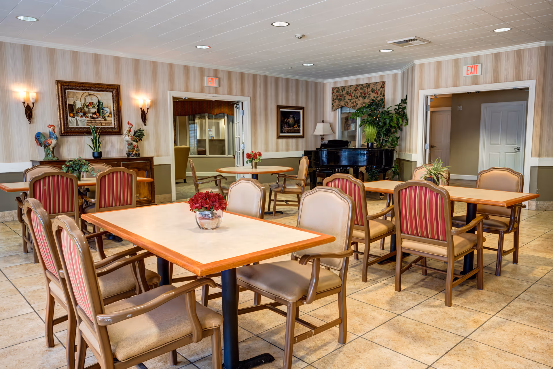 A well-lit dining room with several tables and chairs arranged neatly. The tables have small flower arrangements as centerpieces. The room features beige tiled flooring, striped wallpaper, framed artwork, wall sconces, and a black grand piano in the corner. There are two exit doors visible, and some greenery decorates the space.