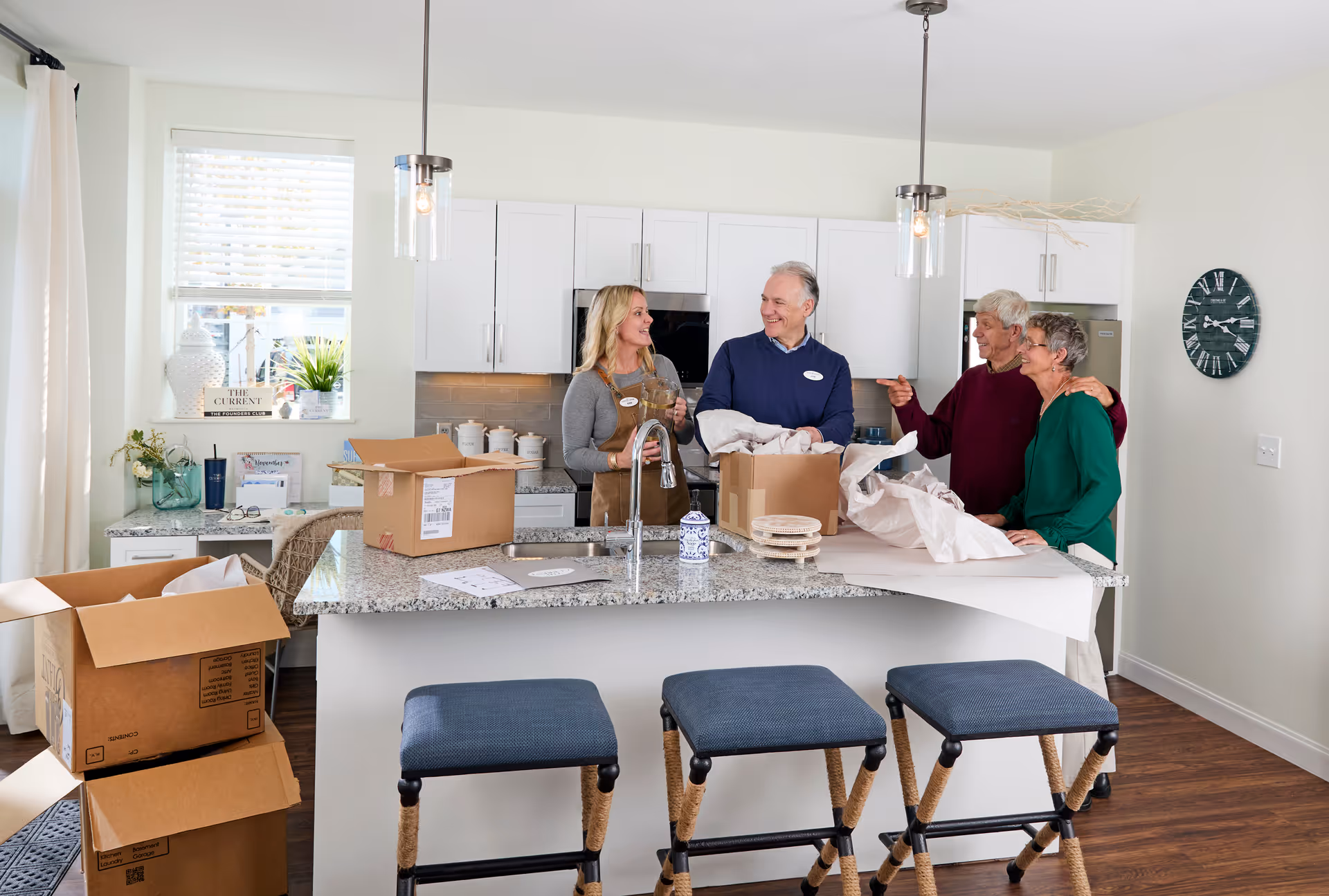 Four older adults standing and talking in a modern kitchen with white cabinets and granite countertops. There are unpacked cardboard boxes on the kitchen island and floor, and three blue cushioned stools in front of the island. A window with blinds and a clock on the wall are visible in the background.