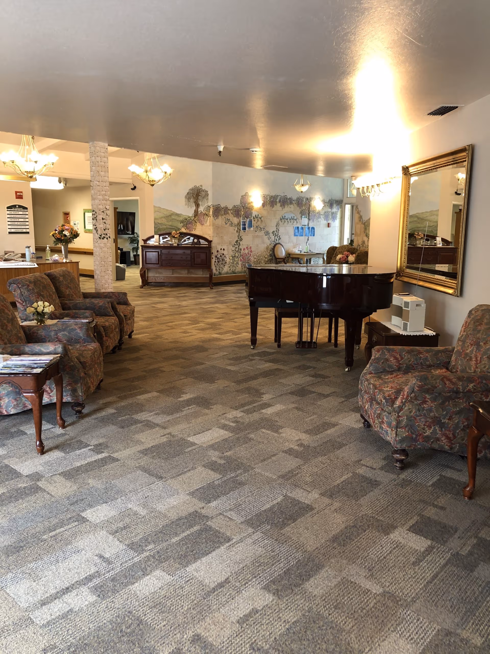 A senior living lounge with patterned armchairs, a grand piano, side tables, and a mural on the far wall.
