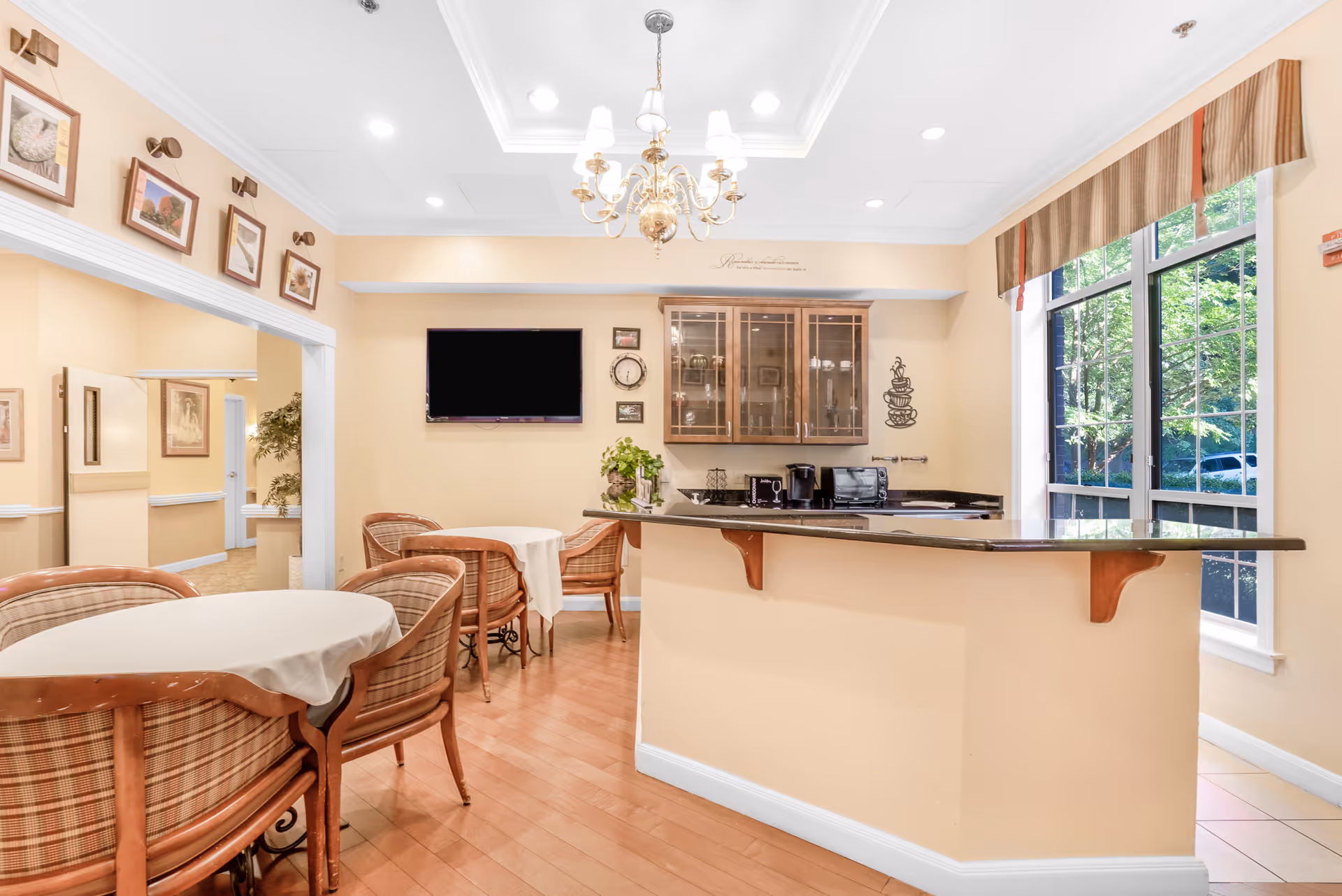 A bright and cozy dining area in a senior living facility featuring round tables with white tablecloths and cushioned chairs. There is a small kitchen counter with a granite countertop, coffee maker, and microwave. The room has light yellow walls, a chandelier, framed pictures on the walls, and a large window with a striped valance letting in natural light.