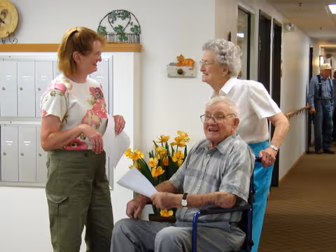 Three elderly individuals in a hallway of a senior living facility. One man is sitting in a wheelchair holding papers, smiling at the camera. Two women stand nearby, one in a floral shirt and green pants holding papers, and the other with white hair wearing a white blouse and blue pants. There are yellow flowers on a table behind them and mailboxes on the wall.
