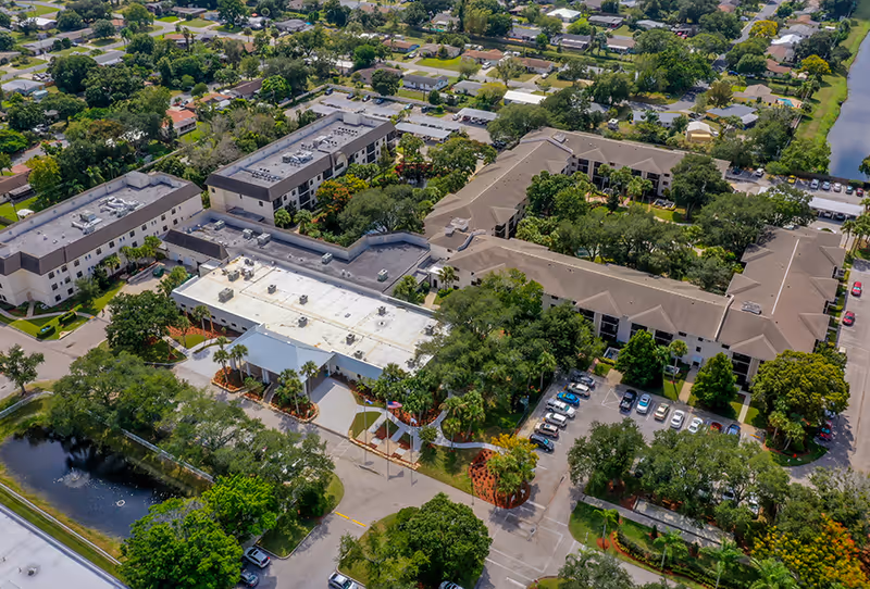 Aerial view of Buena Vida Estates senior living facility showing multiple connected buildings with beige roofs, surrounded by trees, parking lots with cars, and nearby residential neighborhood. There is a small pond with a fountain in the lower left corner and well-maintained landscaping throughout the property.